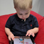 A child sits with a tablet while an adult is nearby, the kind of moment when a parent sets up screen limits.