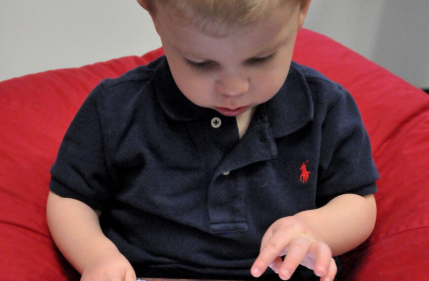 A child sits with a tablet while an adult is nearby, the kind of moment when a parent sets up screen limits.
