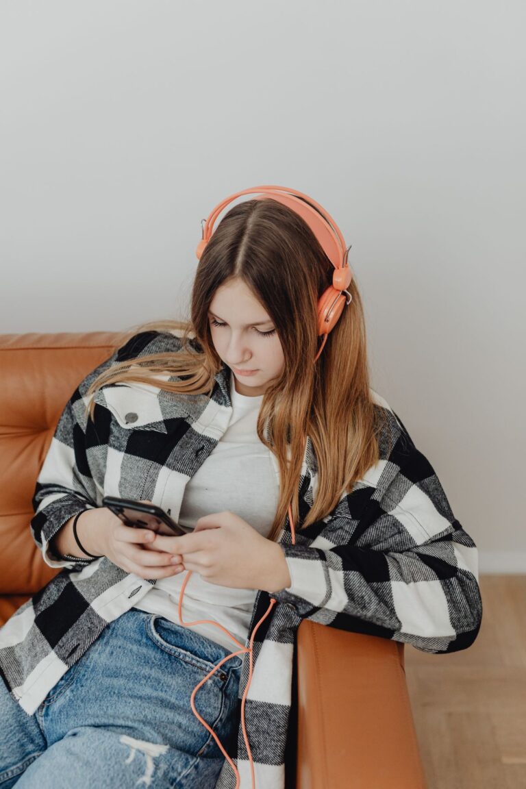 A teenage girl sits on a couch with headphones, looking at a smartphone.