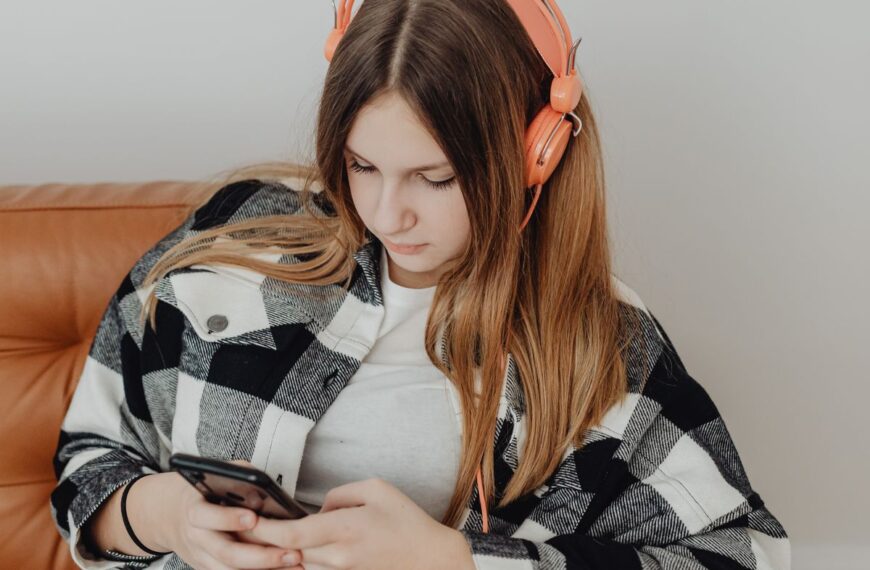 A teenage girl sits on a couch with headphones, looking at a smartphone.